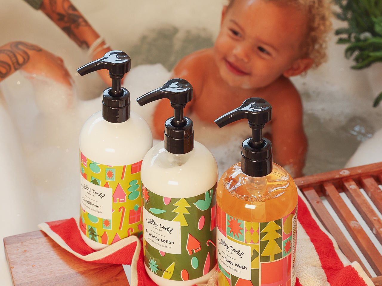 Baby in a bathtub with bubbles, Holiday Collections are in the front (Hair + Body Wash, Conditioner, and Everyday Lotion)